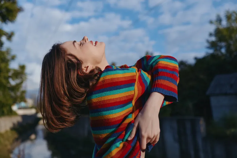 Woman in colorful sweater smiling outdoors in a relaxed portrait, leaning back with eyes closed. Joyful young adult in casual fashion by a riverside under blue sky.