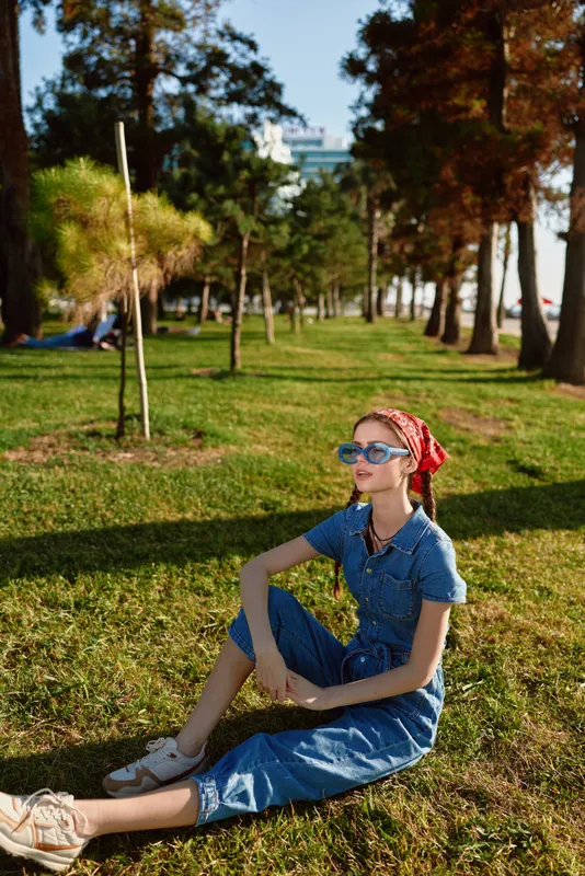 Lifestyle woman in modern boho-western denim street style sitting outdoors on grass with sunglasses and red bandana, warm film color creating relaxed atmosphere.