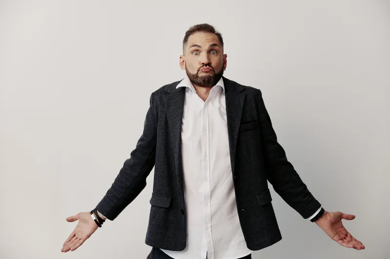 A professional man in a blazer stands on a colored isolated background, presenting a curious pose with open arms and a subtle hint of uncertainty.