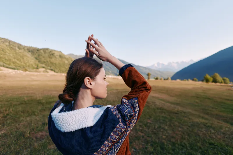 Woman in nature standing in a meadow with mountains ahead, outdoors portrait from behind wearing patterned jacket, peaceful landscape and open field at golden hour soft natural light.