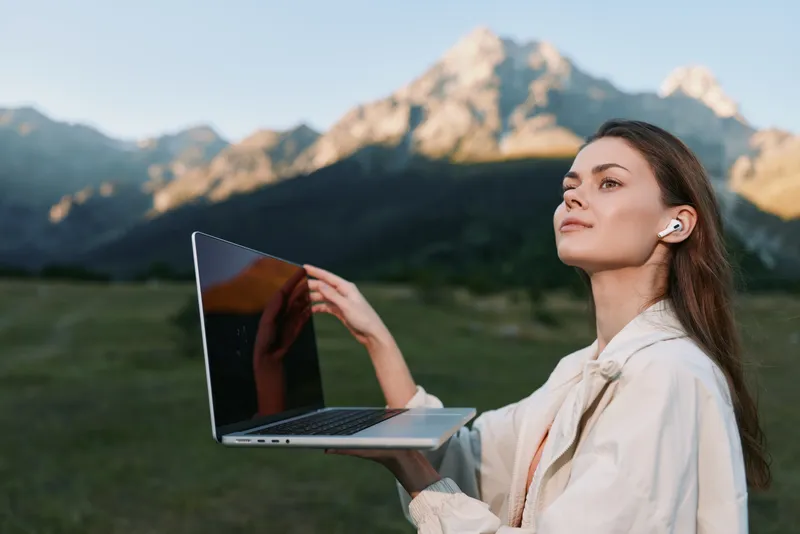 A confident outdoor professional uses a laptop in a scenic landscape, communicating clearly as majestic mountains rise behind, creating a calm and productive atmosphere.
