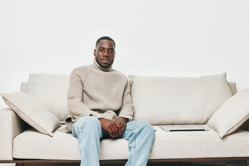 A young African man sitting on a light colored sofa, wearing a cozy turtleneck sweater and jeans, exuding a calm emotion in a minimalist living room