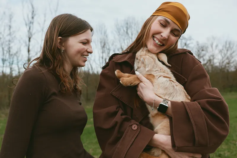 Two women outdoors share a joyful moment with a small goat kid, cuddling and smiling. Cozy outfits and a natural landscape convey friendship, warmth, and rural lifestyle on a breezy day in a field