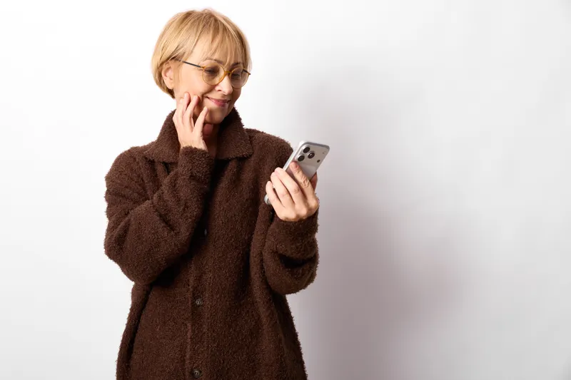 Smiling elderly woman in brown coat portrait holding smartphone device, messaging communication concept, warm indoor lighting, white studio background