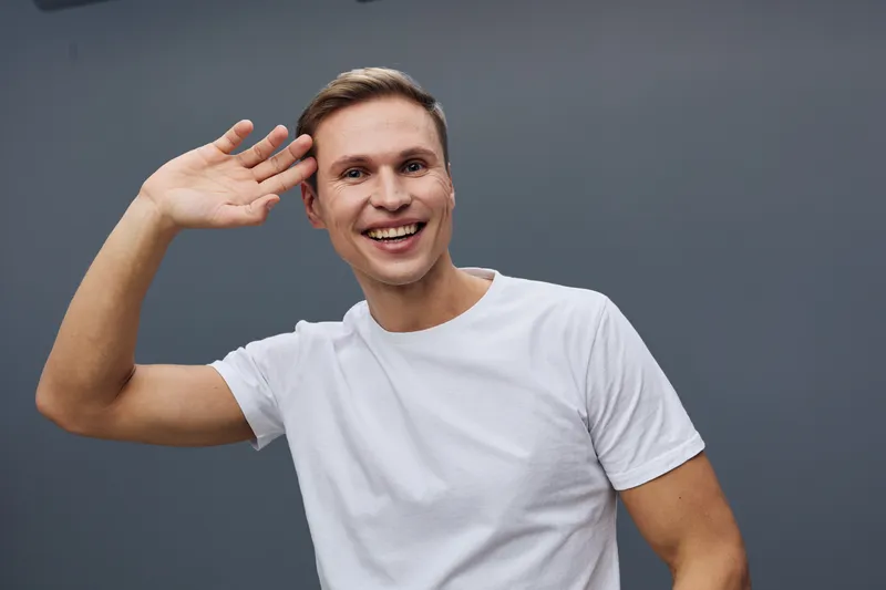 Smiling man with short brown hair wearing white t-shirt salutes with hand near head, looking cheerful and friendly, isolated on gray background, studio portrait, positive emotions, casual style, happi