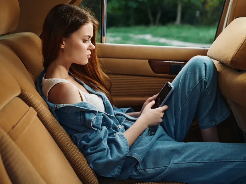 Young woman in casual denim outfit sitting in a car, absorbed in her smartphone. Natural light highlights her pensive expression and relaxed posture.