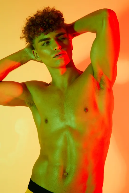 Young man with curly hair posing shirtless with hands behind head, showing muscular torso and defined abs under colorful green and orange lighting in studio setting.
