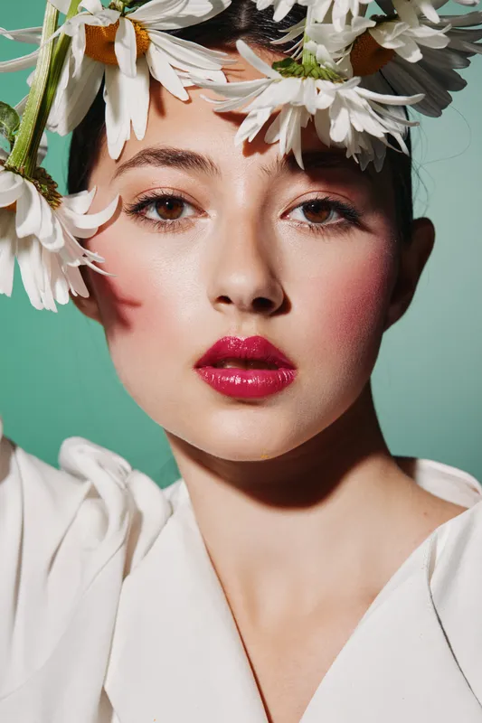 Closeup of young woman with creative floral headpiece wearing glossy pink makeup, soft skin, and styled outfit. Minimalistic background highlights experimental fashion beauty concept.
