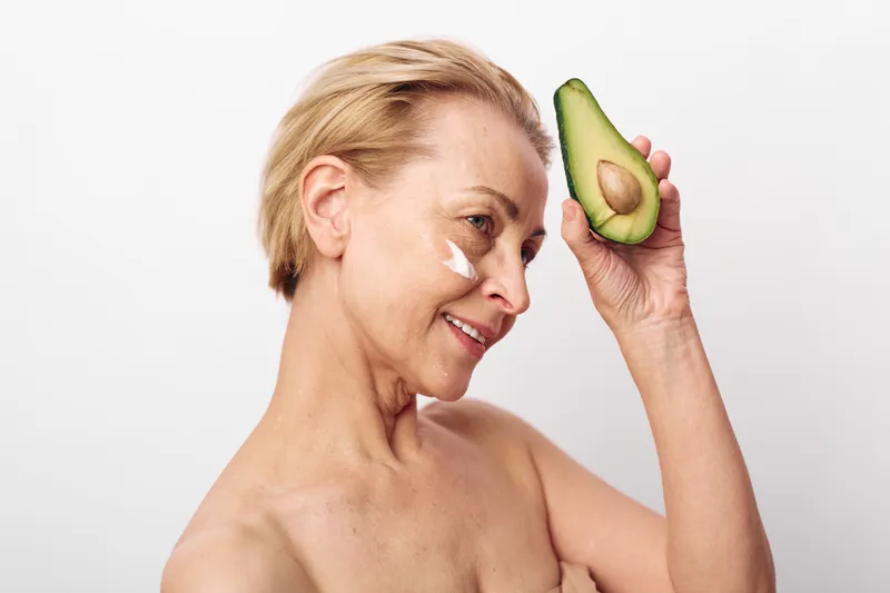 Mature woman with short blonde hair applies natural cream on face holding avocado half near head isolated on white background. Concept of skincare, healthy lifestyle, natural beauty and wellness.
