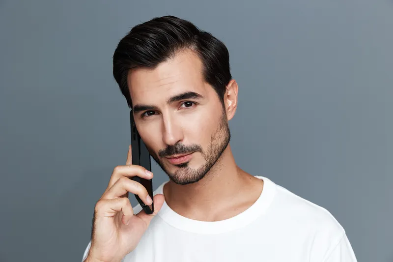 Handsome man with a stubbled beard holding smartphone near ear, looking confidently at the camera with a neutral gray background, casual white shirt attire.