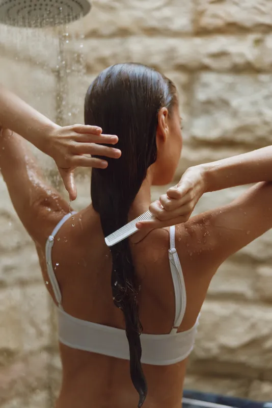 Woman combing wet hair under warm outdoor shower with stone wall background, natural light enhancing smooth skin and relaxed summer atmosphere.