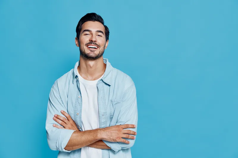Smiling man wearing a light blue shirt stands with crossed arms against a vibrant blue background, radiating joy and confidence in a studio setting.