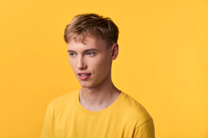 Young man wearing a yellow shirt stands against a bright yellow background in a studio shot, showcasing a casual pose, subtle smile, and modern fashion style with clean composition.