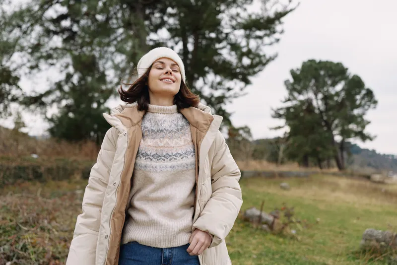 Outdoor woman wearing a cozy sweater and coat, smiling in a breezy countryside field with trees, capturing a relaxed casual fashion moment for lifestyle photography.
