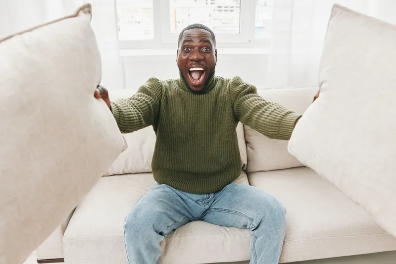 Happy young Black man expressing joy while sitting on a sofa, holding decorative pillows in a cozy living room setting with soft lighting