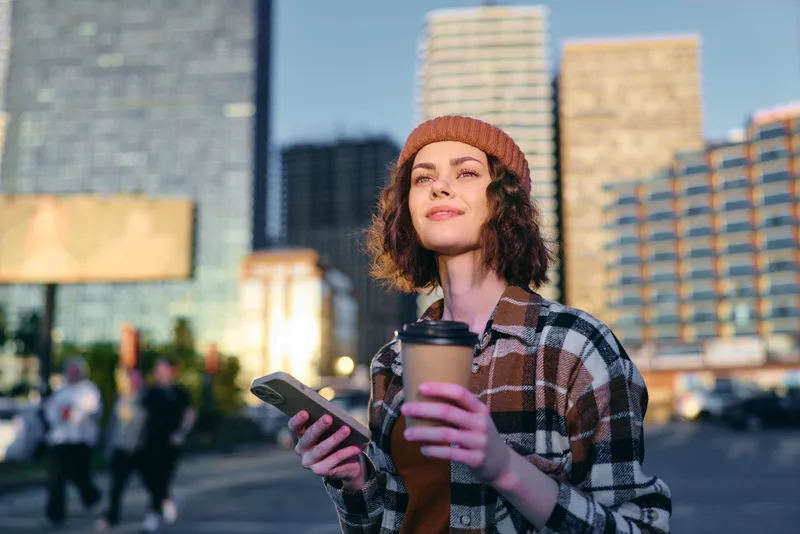 woman holding coffee and smartphone in an urban street at golden hour, candid lifestyle shot conveying authenticity, mindful living and emotional storytelling with warm glow.
