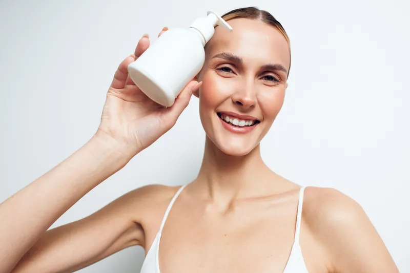 Smiling young woman applies skincare from a white bottle in a bright studio, enjoying a fresh beauty moment and relaxed vibe.