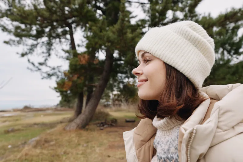 Woman in a warm coat and cream knit hat gazes toward a misty outdoor landscape with trees and open field, serene side profile, soft natural light, calm moment captured in nature.