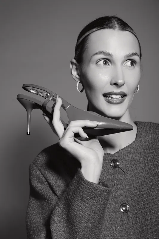 Portrait of a fair skinned caucasian woman holding a high heel shoe near her face, wearing a textured coat and hoop earrings, looking aside with a curious expression.