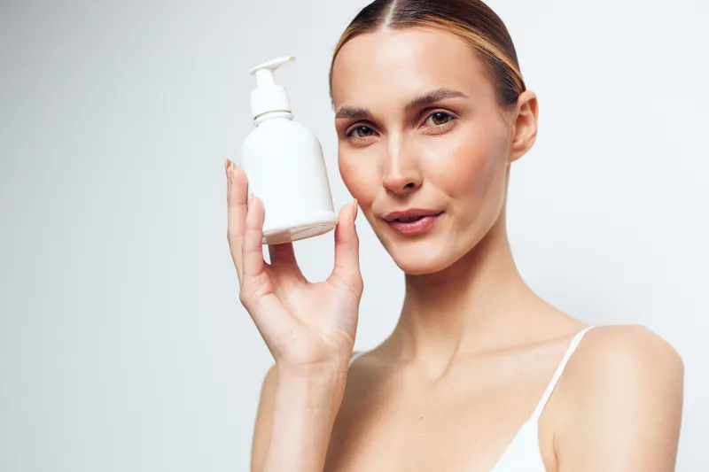 Young woman with clear skin holding a white lotion bottle close to her face. Minimalist background emphasizes skincare, beauty, and personal care concepts with soft natural lighting.