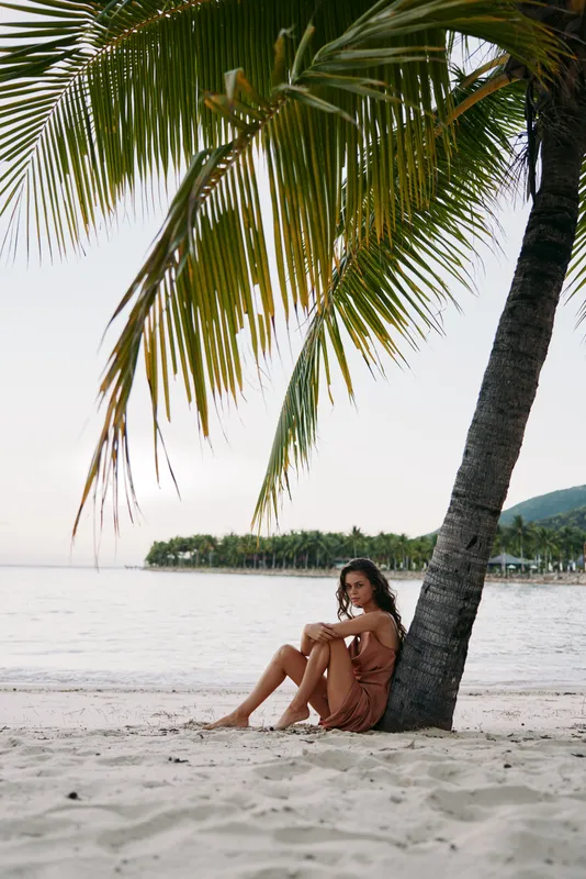 Young woman sitting by the palm tree on the beach at sunset, relaxed and thoughtful with tropical background and calm sea.