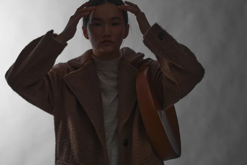asian woman wearing a warm brown coat stands in a dim setting, hands raised near her head, moody lighting highlights texture and evokes a thoughtful, fashion oriented moment.