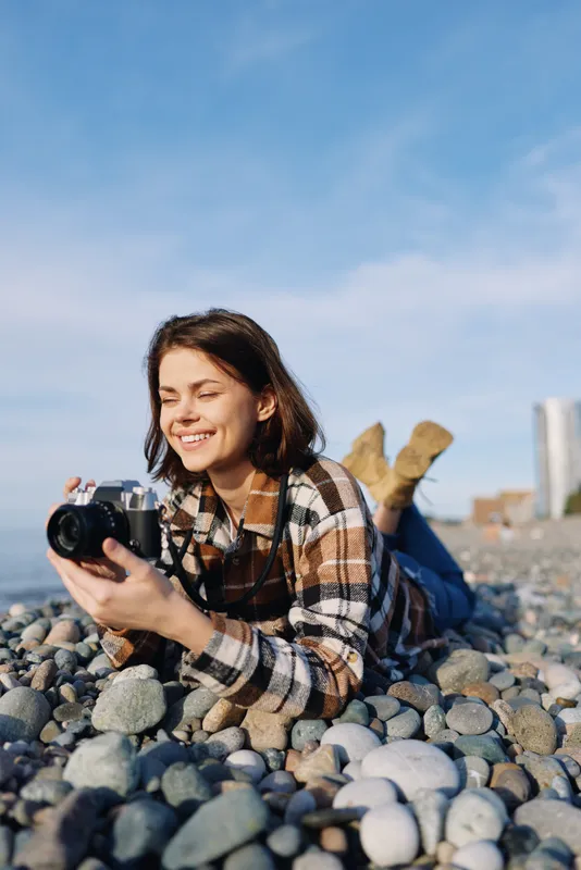 Young photographer lying on a rocky beach, smiling while holding a camera, outdoor scene under clear sky, plaid shirt, natural light, candid storytelling and creative exploration through photography