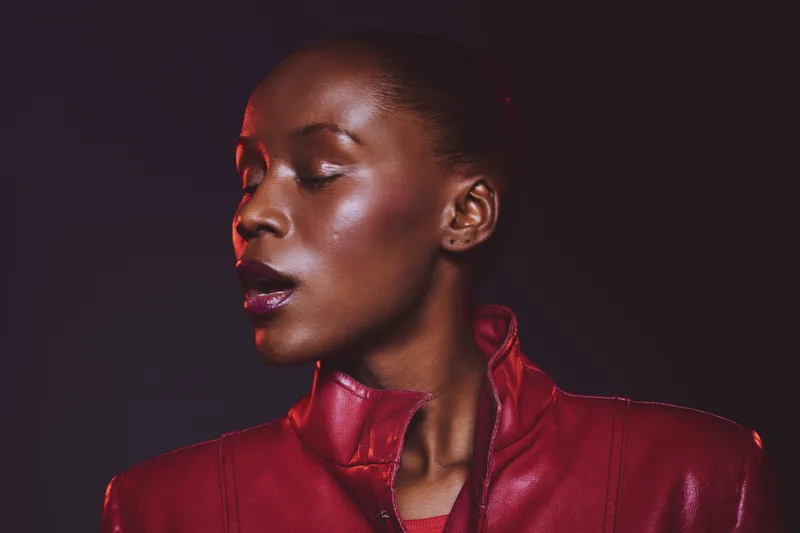 Close-up of an elegant African woman with dark skin wearing a red leather jacket, eyes closed against dark background. Concept of fashion, beauty, and confidence in studio lighting.