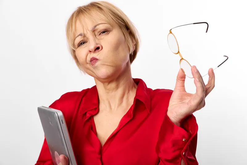 Senior woman in red blouse looks surprised while holding eyeglasses and a phone, concerned about vision and eyesight, concept of eye health and aging care today