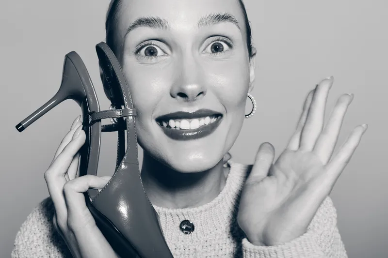 Smiling white woman with light skin holding red high heel shoe near face waving hand with excitement on plain background.