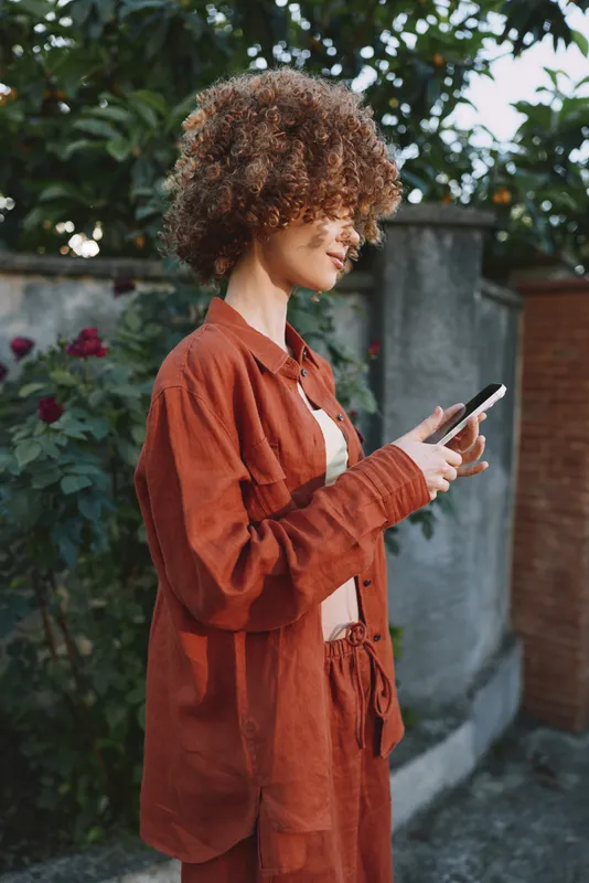 Woman lifestyle portrait outdoors in orange suit uses a smartphone, relaxed pose, casual fashion, natural light, urban garden setting, candid moment, contemporary style, confident demeanor