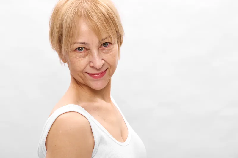 Woman portrait senior blonde smile studio white headshot short hair, soft light, relaxed posture, gentle expression, clean background, closeup of face, and shoulders.