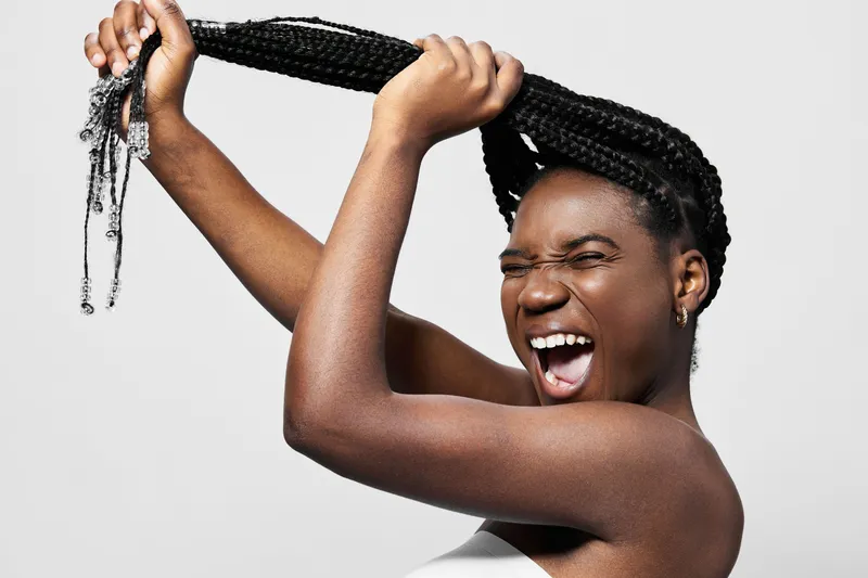Young woman with braids joyfully pulling her hair, showcasing vibrant emotions and a playful expression against a plain background, representing confidence and self expression