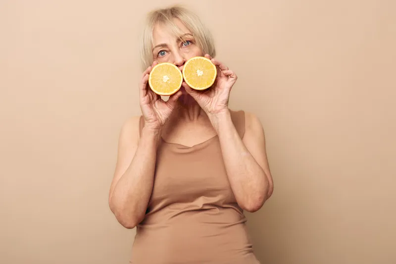 Middle aged woman with blonde hair holds two halves of grapefruit near face, wearing beige top against beige background. Concept of healthy eating, vitamin C, and natural lifestyle.