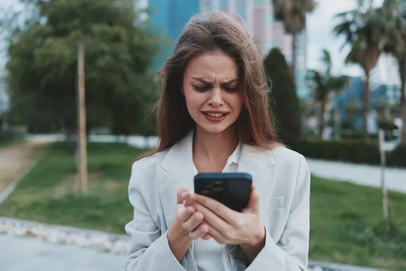 Woman stressed by financial crisis holding smartphone outdoors in casual business attire with urban background and emotional expression