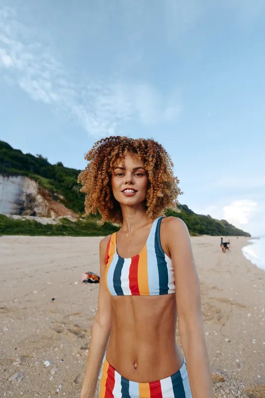 Woman on beach with curly hair, wearing striped swimsuit, smiling in sunlight, outdoor summer walk, gentle sea breeze, vibrant colors, sandy shoreline and green cliffs background clear sky