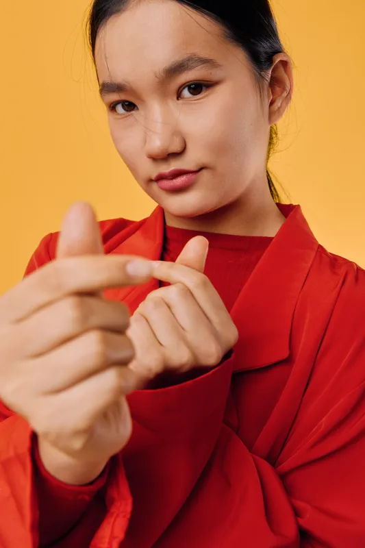 Woman in red jacket and shirt shows hand gesture with fingers on yellow background. Portrait of confident female with tied hair and natural makeup in casual clothing for communication.