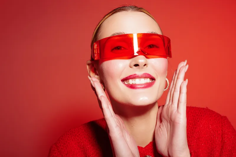 Smiling woman in red sweater and futuristic red sunglasses poses against red background. Bright studio lighting highlights her joyful expression and modern fashion style.