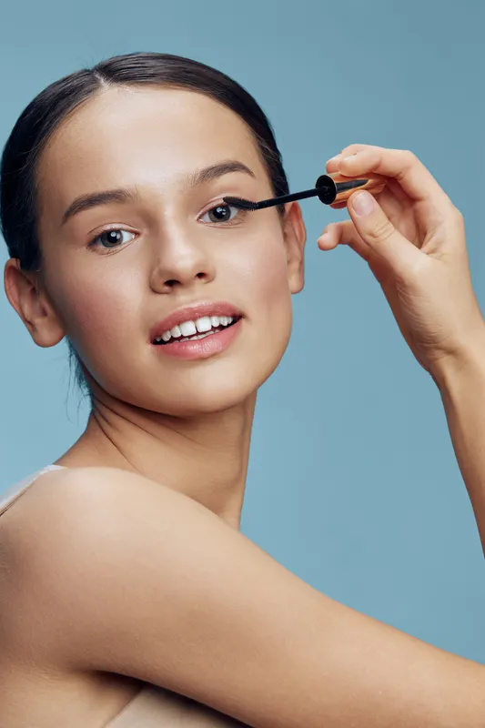 Young woman applying mascara with a bright smile, showcasing beauty and confidence against a soft blue backdrop, emphasizing makeup and self care