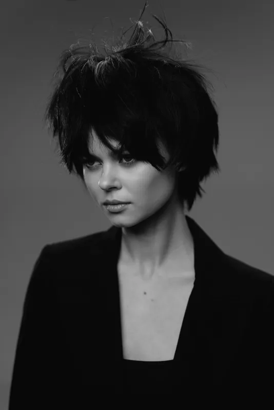 Black and white portrait of a stylish woman with short hair and serious expression in a dark outfit and neutral background.