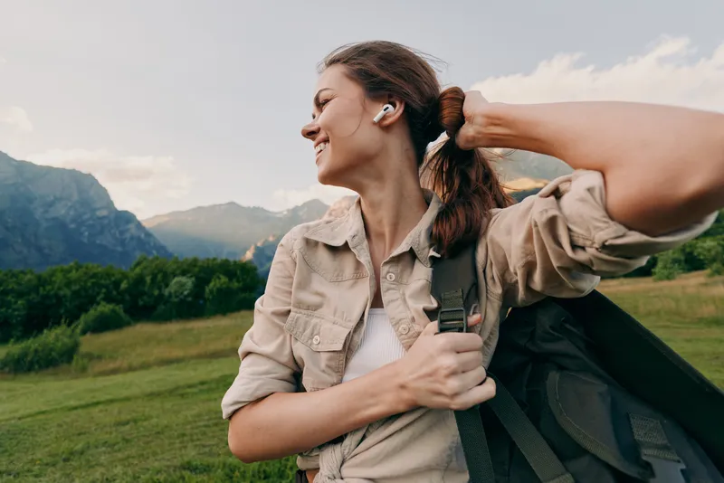 Young woman with curly hair adjusts her ponytail while hiking outdoors in a beige jacket and white t-shirt with mountains and green fields in the background.