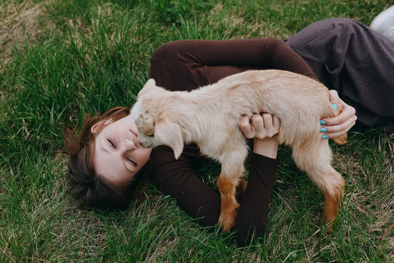 Young woman lies on grass holding a baby goat close to her face in a peaceful outdoor lifestyle moment with natural light and casual clothing