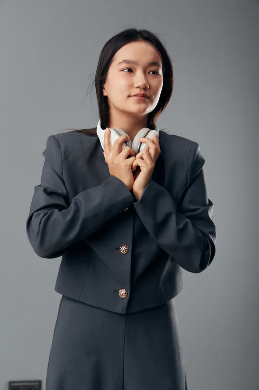 Young woman in gray suit holding white headphones standing against gray background looking aside with thoughtful expression. Professional female portrait with casual technology accessory.