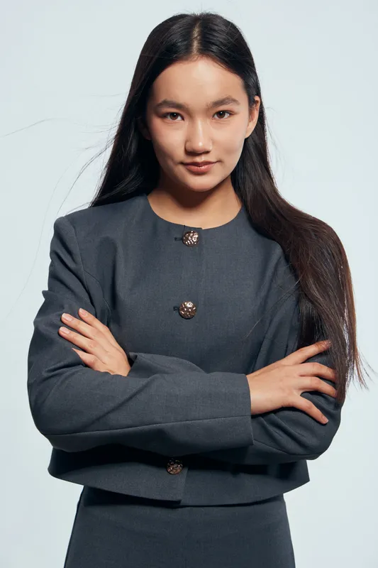 Portrait of confident young woman with long dark hair wearing dark gray blazer standing with crossed arms looking at camera with slight smile on light background.