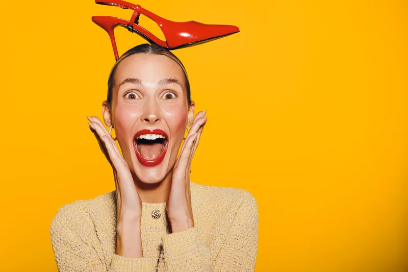 Excited woman with red lipstick and beige sweater holds red high heel shoe on head against orange background. Concept of fun, fashion, and playful mood with bright studio lighting.
