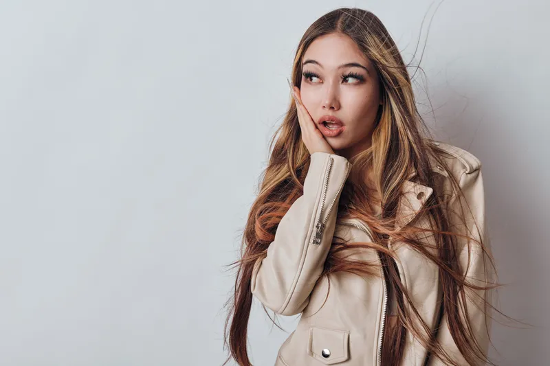 Woman with long hair and stylish jacket posing with hand on face looking away, expression of surprise or contemplation on light background in studio portrait.