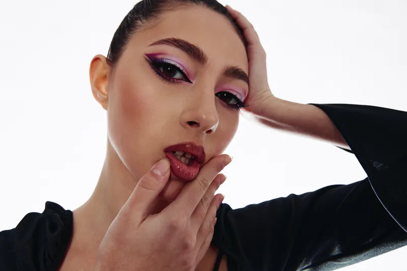 Young woman with bold pink eye makeup and glossy lips poses expressing elegance and confidence. Studio shot on a clean white background with creative beauty and fashion concept.