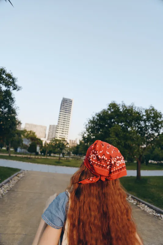 woman with red hair wearing a red bandana walking in urban park under clear sky with green trees and modern buildings in background during daytime