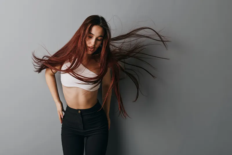 Woman with long hair in motion wearing white crop top and black pants against gray background. Casual style, movement, fashion and youth with natural look and confident pose.