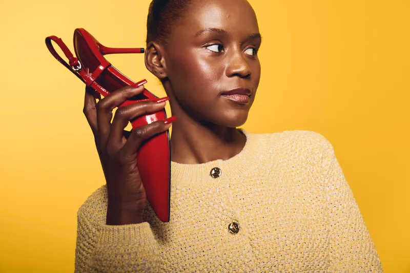 Portrait of a dark skinned African woman holding a red high heel shoe near her ear, wearing a beige knitted sweater against a yellow background. Concept of fashion and style.
